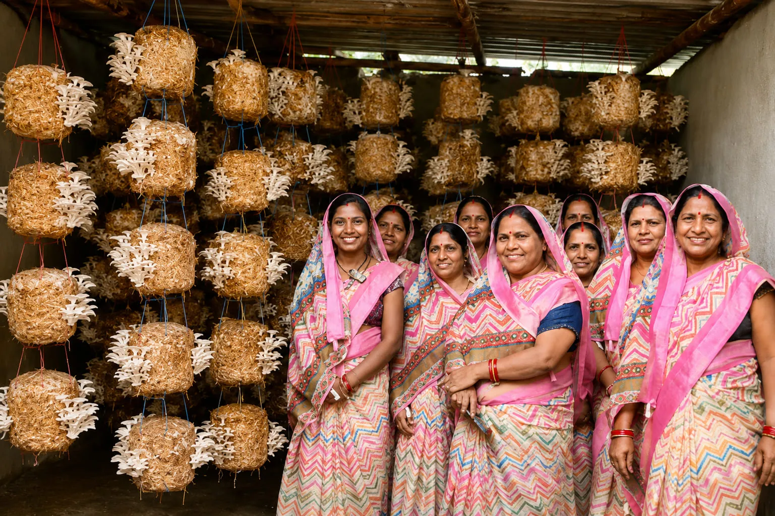 The ten members of Taranga Self-Help Group standing among the hanging mushroom beds in their cultivation shed